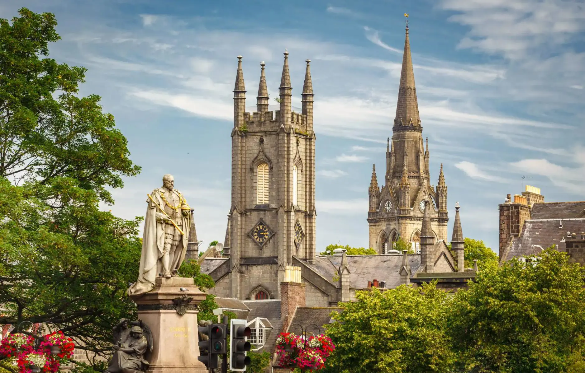 Statue of a historical figure in front of two tall Gothic-style church towers in Aberdeen, surrounded by green trees and hanging flower baskets under a partly cloudy sky