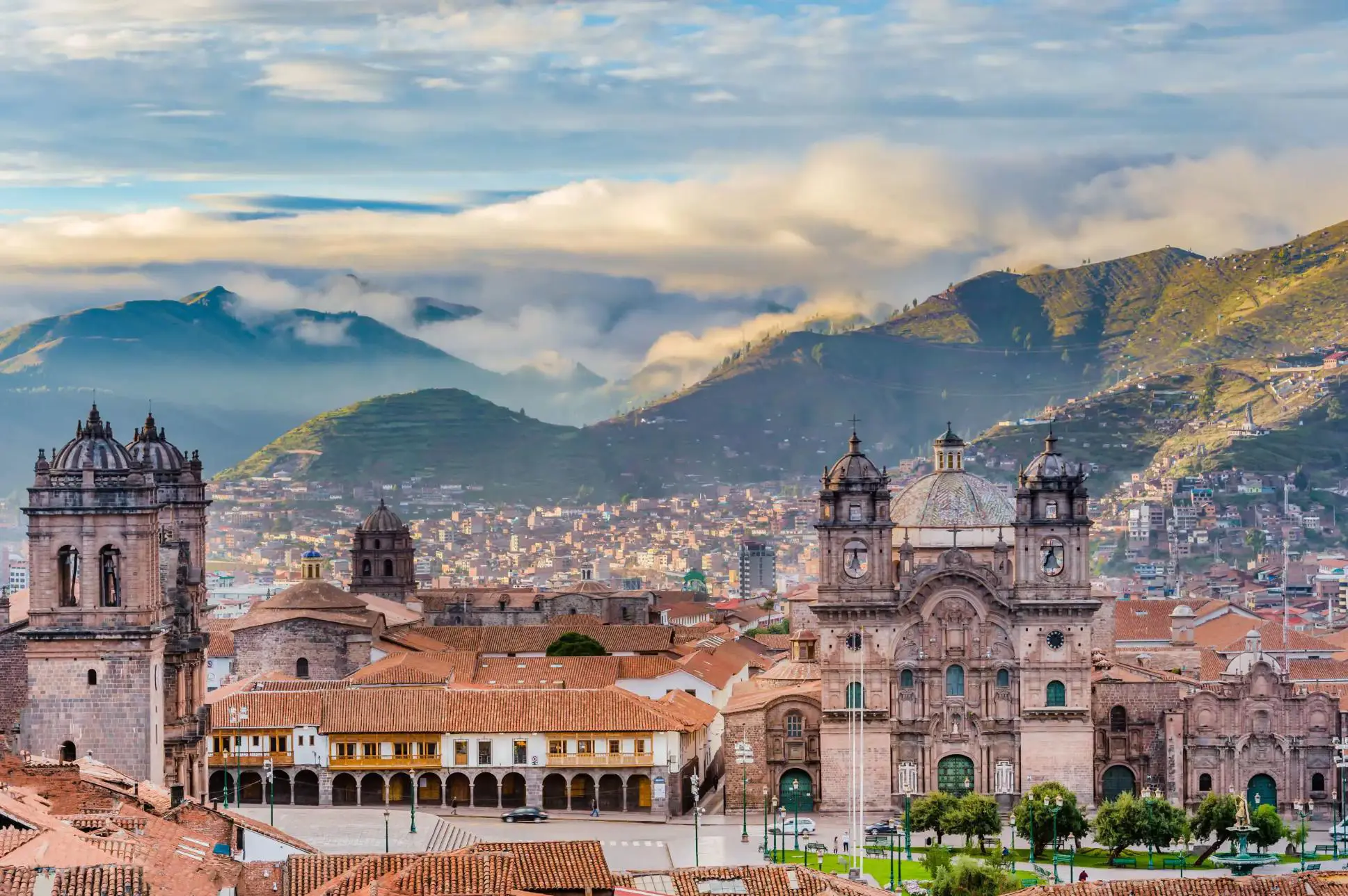 A panoramic view of Cusco, Peru, showcasing historic colonial buildings with terracotta roofs against a backdrop of green mountains and a cloudy sky.A panoramic view of Cusco, Peru, showcasing historic colonial buildings with terracotta roofs against a backdrop of green mountains and a cloudy sky