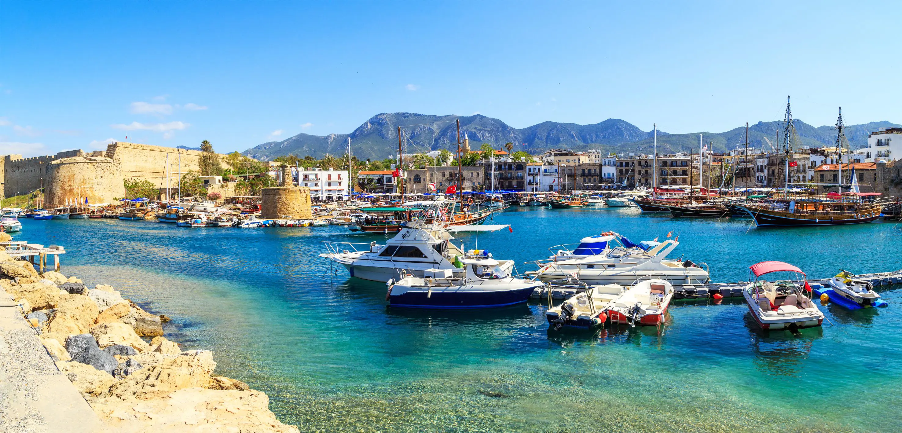 Boats docked at a harbour with bright blue water, buildings on the other side, and ruins of a castle to the left. Mountains in the distance in front of a blue sky.