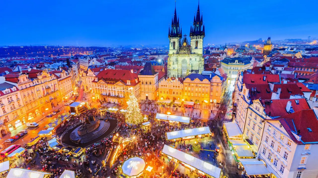 High angle shot of a Christmas market in the town square of Prague, showing a lit up gothic church with two towers and a large lit up Christmas tree