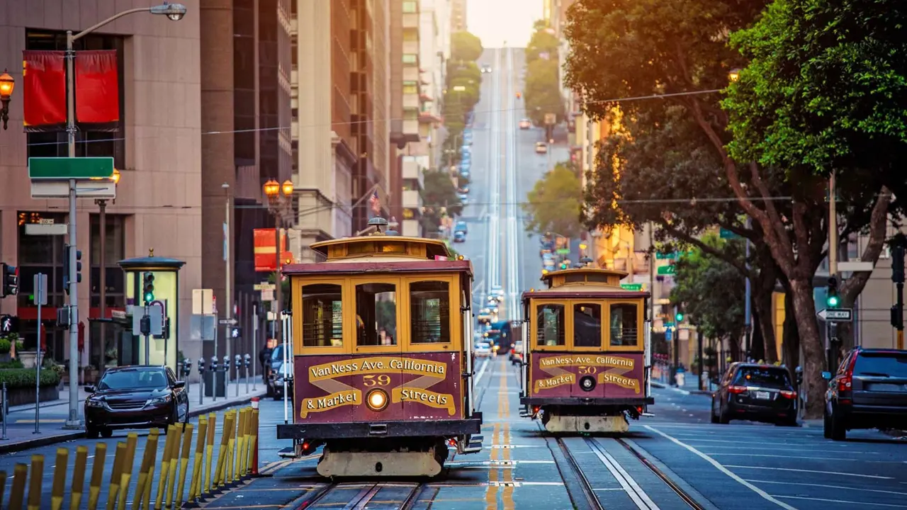 Two traditional San Francisco cable cars in yellow and maroon colours descending a steep hill with parked cars either side of the street